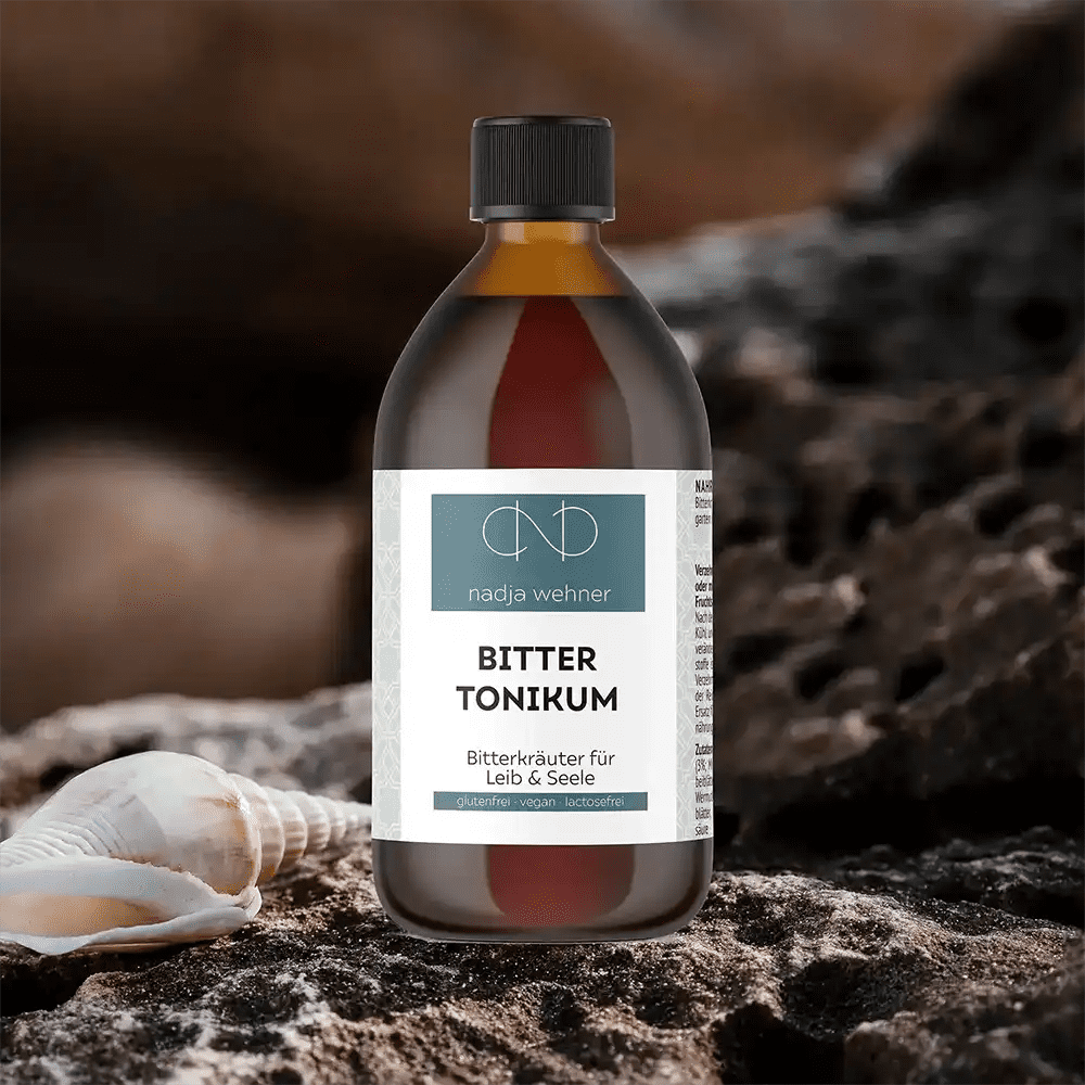 Eine Flasche Bitter Tonikum der Rosen Apotheke Manufaktur mit einer dunklen Flüssigkeit steht auf felsigem Gelände neben Muscheln und fängt die Essenz des Bitterkrauts ein.